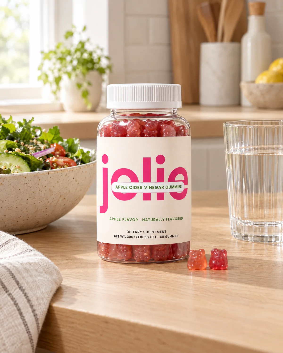 A jar of jolie Apple Cider Vinegar gummies on a kitchen counter next to a salad and a glass of water